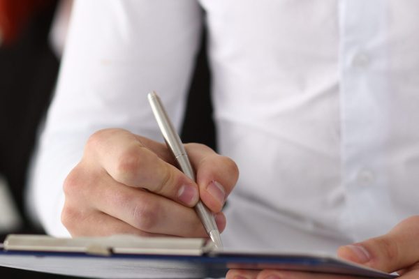 Woman and man are using laptop jointly while male is sitting on desk and writing information on paper